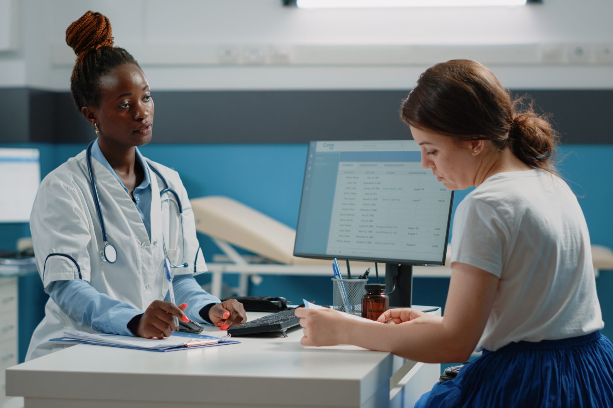 Dos mujeres una médica y otra paciente en un consultorio médico revisan un documento, al fondo una camilla