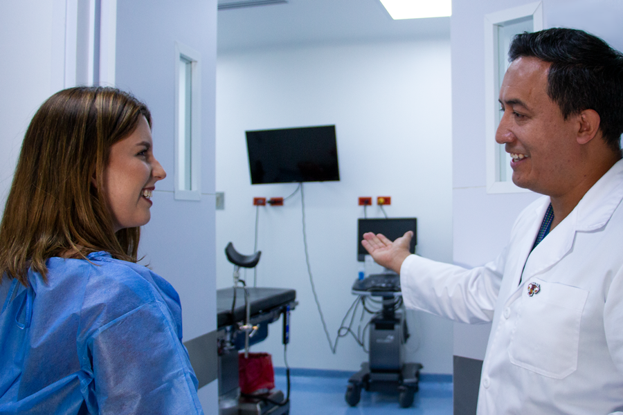 Dos mujeres una médica y otra paciente en un consultorio médico revisan un documento, al fondo una camilla